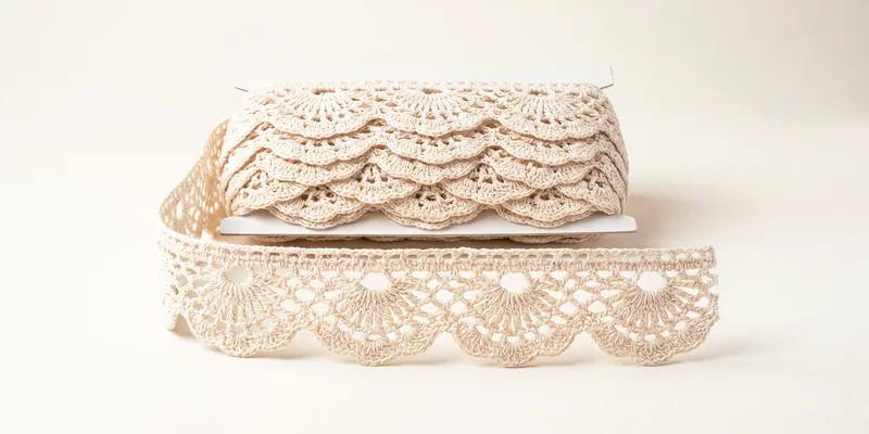 A flat card wound with cream scalloped crochet lace, a length unwound in front lying flat to show the fan motifs and open holes along the scalloped edge