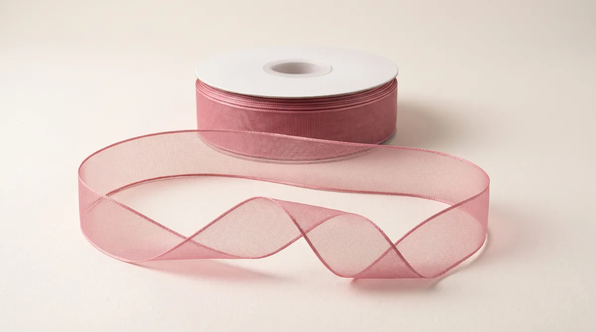 A reel of dusty rose organza ribbon with a length unwound in front, the sheer weave showing the surface beneath it where the ribbon twists
