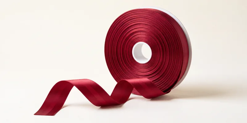 A roll of burgundy satin ribbon on a white core standing on edge, with a length unwound in front curling to show the glossy face