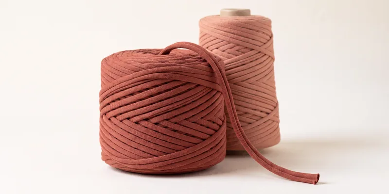 Two put-ups of terracotta t-shirt yarn, a flat cake in front and a cone behind, with a length of the flat ribbon yarn draped across and its cut end showing