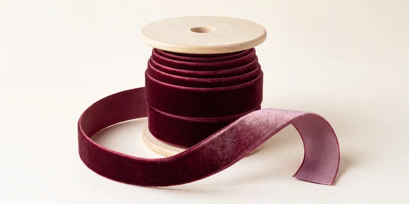 A wooden spool of burgundy velvet ribbon with a length unwound into a loop, showing the soft pile face and, where the ribbon twists at the end, the paler satin reverse